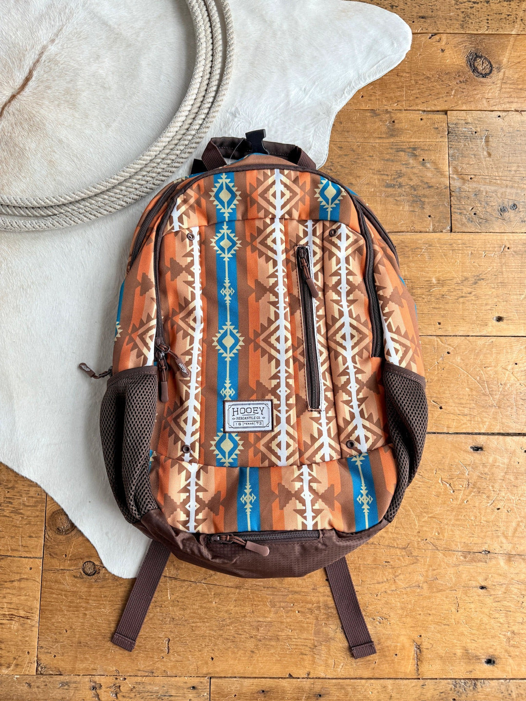 The Calexico Backpack by Savannah Sevens western life{&}style, featuring a brown aztec print with tan, white, and blue geometric patterns and a padded laptop compartment, sits on a wooden floor near a coiled rope and white cowhide rug. By Hooey. 