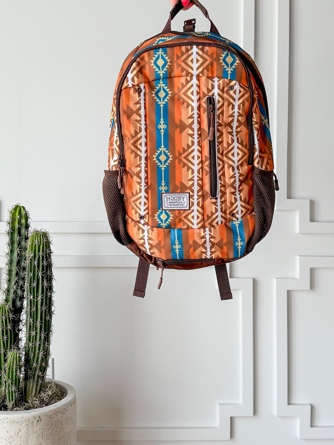 The Calexico Backpack by Savannah Sevens western life{&}style, featuring an Aztec print and padded laptop compartment, hangs on a white paneled wall next to a potted cactus. By Hooey. 
