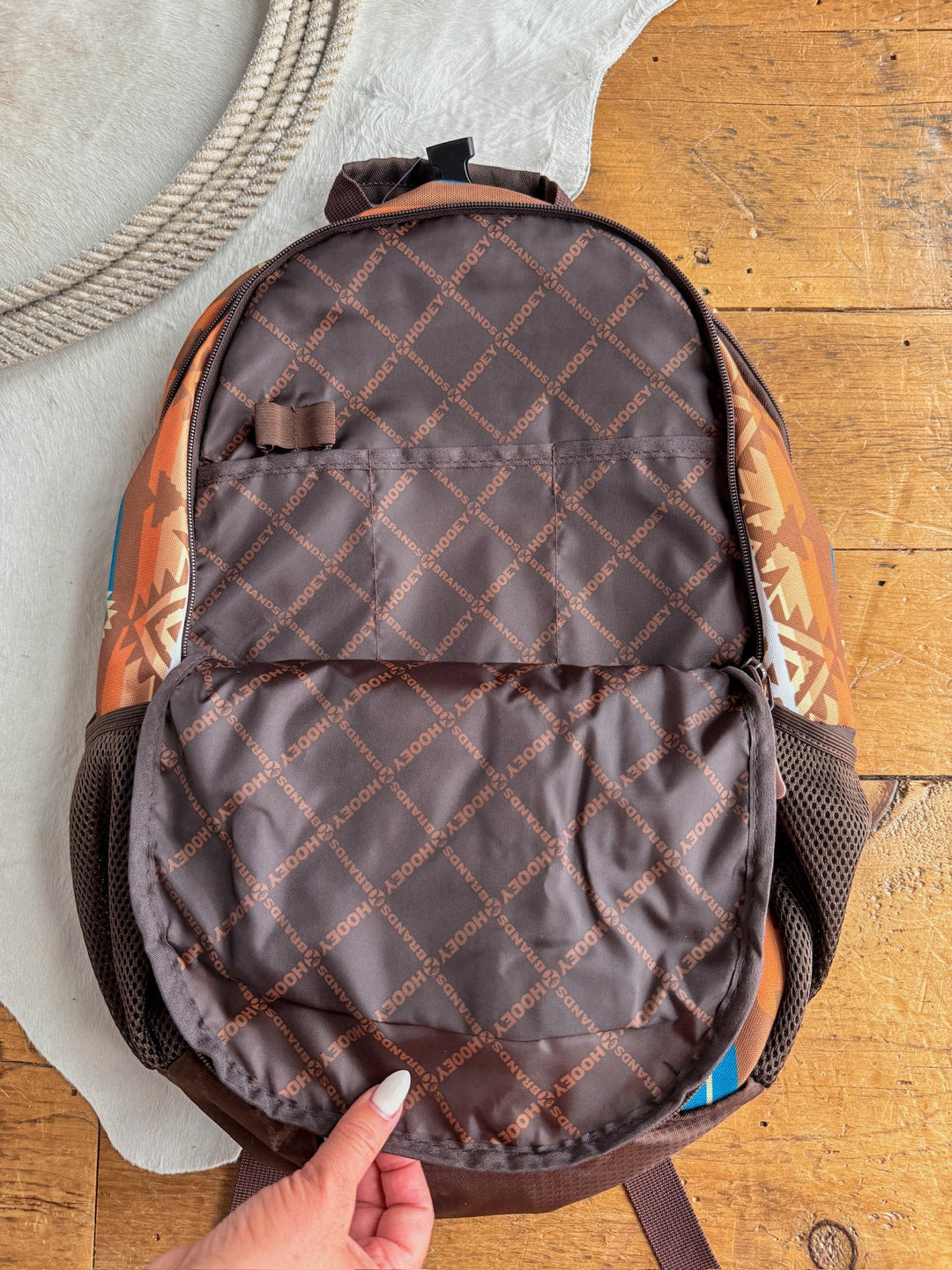 A hand opens the Calexico Backpack by Savannah Sevens western life{&}style, showing its branded interior. The brown Aztec print bag rests on wood near a white rug and rope, and features a padded laptop compartment for functionality. By Hooey. 