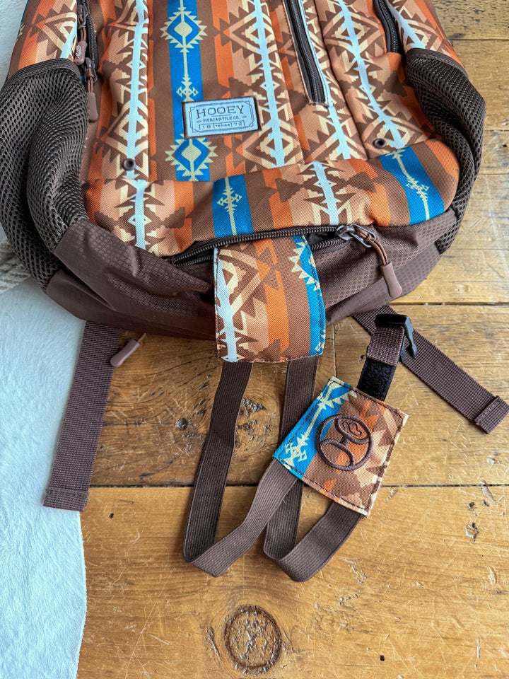 The Calexico Backpack by Savannah Sevens western life{&}style features a brown and orange Aztec print, blue accents, mesh side pockets, padded laptop compartment, HOOEY patch, adjustable straps, and a keychain with a stylized logo.