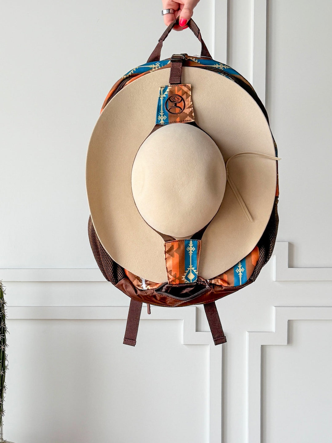A hand holds the Savannah Sevens western life{&}style Calexico Backpack with an Aztec print and a beige wide-brimmed hat attached to the front, featuring a padded laptop compartment, set against a white paneled wall. By Hooey. 