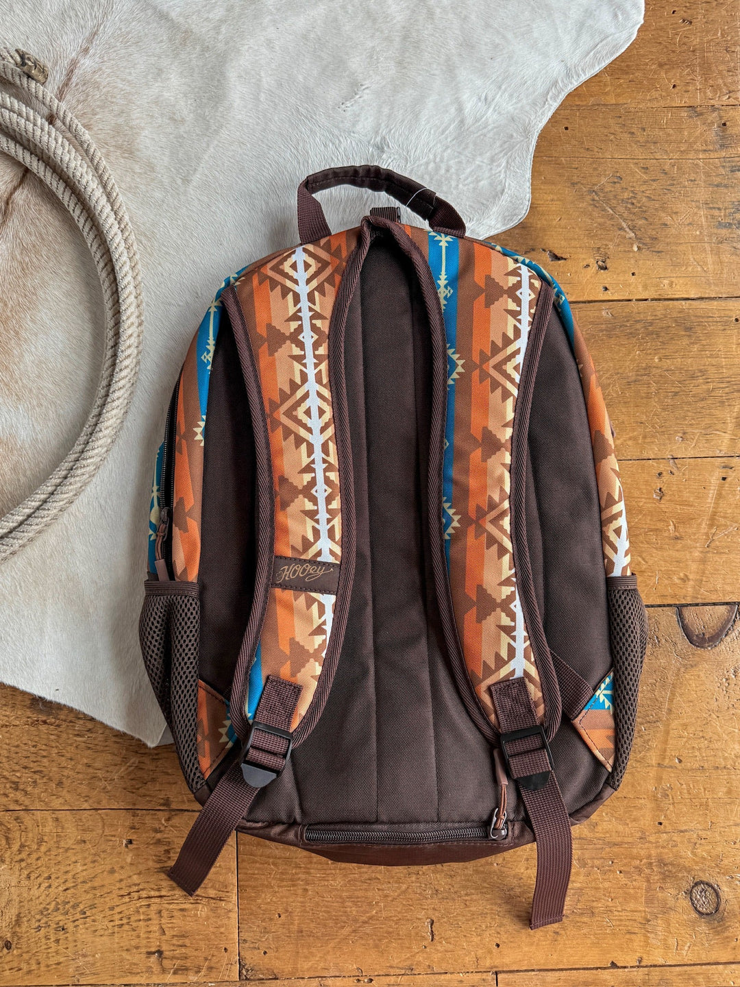 The Calexico Backpack by Savannah Sevens western life{&}style, with brown and orange Aztec geometric designs and a padded laptop compartment, sits on a wooden floor beside a white rug and coiled rope. By Hooey. 