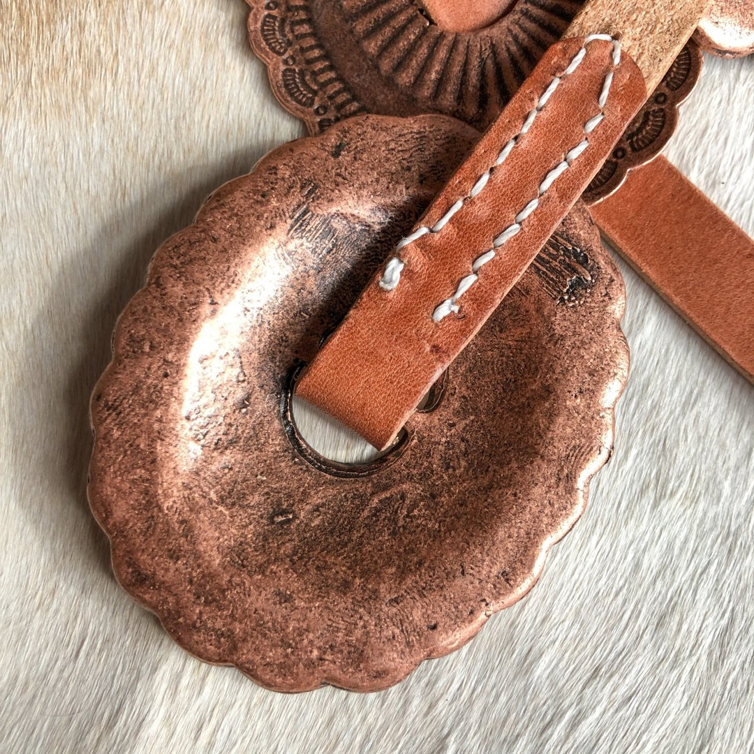 Close-up of the Calexico Belt by Savannah Sevens western life{&}style, featuring a decorative round copper concho with a scalloped edge on distressed harness leather with white stitching, displayed on a tan, fur-like surface.