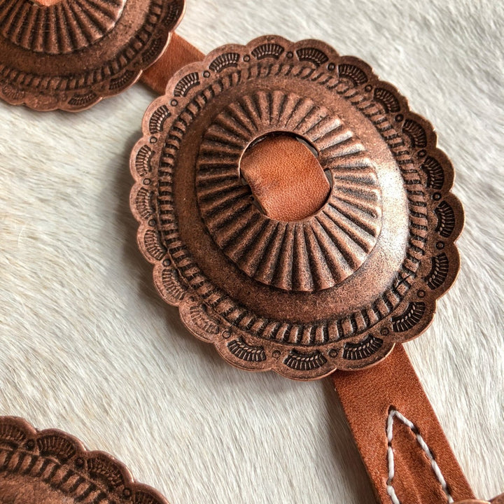 A close-up of the Calexico Belt by Savannah Sevens western life{&}style shows an ornate round copper concho with a scalloped edge and engraved details on distressed harness leather, highlighted by visible stitching against a light textured background.