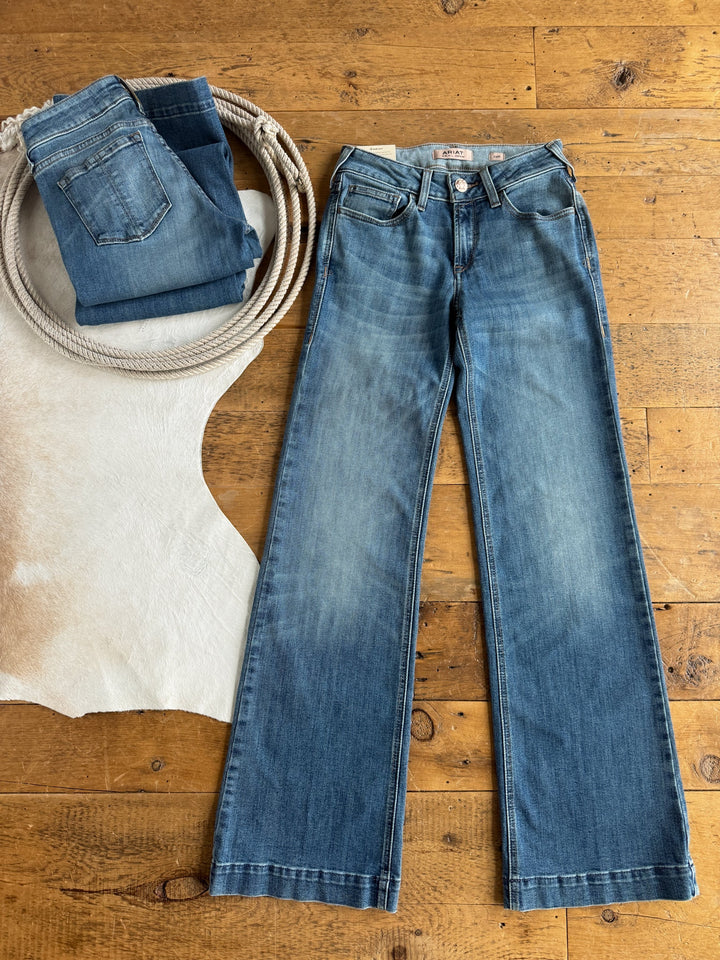 Two pairs of the Callahan Perfect Rise Trouser Jeans by Savannah Sevens western life{&}style—one folded and one laid flat—rest on a wooden floor, accompanied by a coiled rope and a glimpse of a white cowhide rug. These exclusive jeans feature classic medium wash denim, delivering timeless style and versatility.