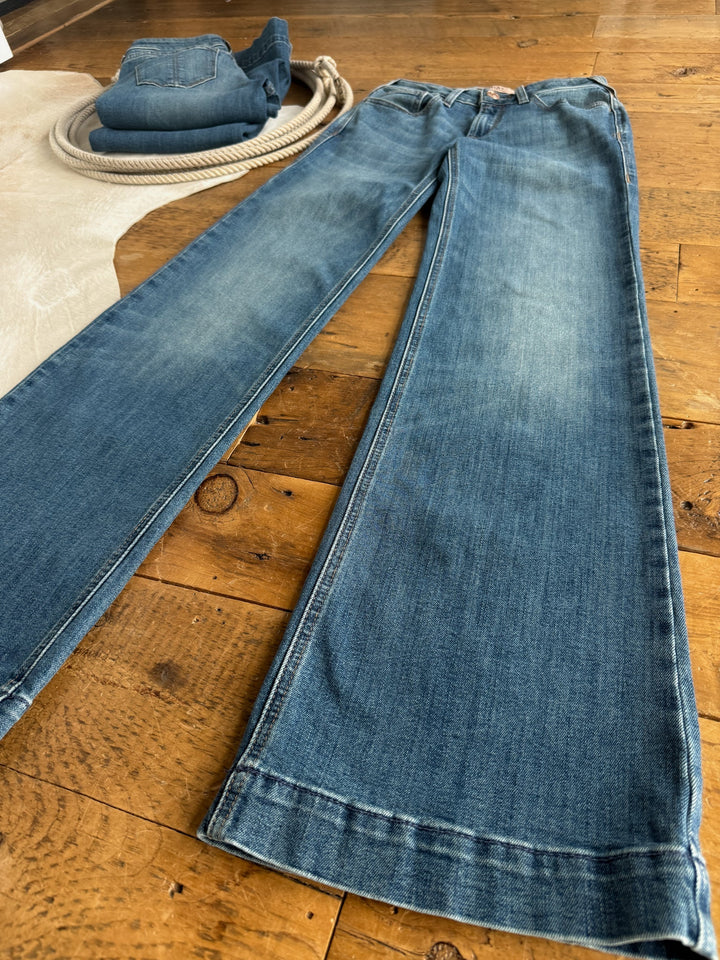 The Callahan Perfect Rise Trouser Jean by Savannah Sevens western life{&}style is laid flat on a wooden floor, with another folded pair of jeans and a coiled rope in the background.