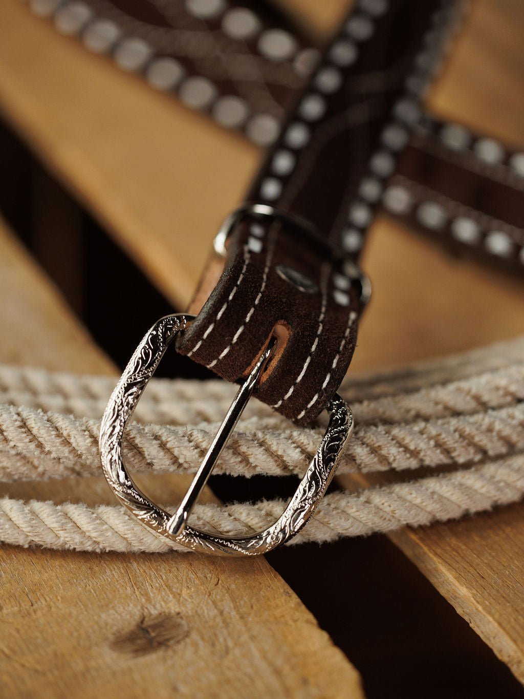 A close-up of the Camargo Belt by Savannah Sevens western life{&}style, featuring brown rough out leather, white stitching, and an ornate sterling silver buckle, displayed on a light rope and wooden background.