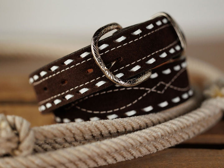 The Camargo Belt by Savannah Sevens western life{&}style features dark brown rough out leather with white stitching and an engraved sterling silver buckle, shown coiled on wood next to a looped rope.