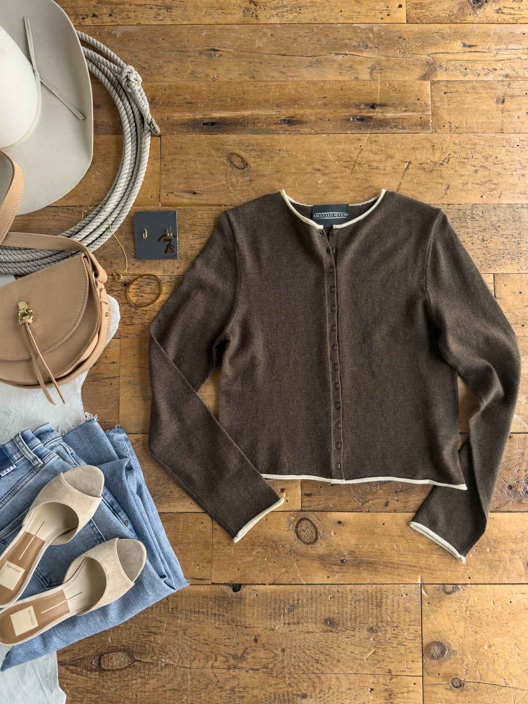 The Camargo Brown Button Up Cardigan from Savannah Sevens western life{&}style is displayed in a flat lay on a wooden floor alongside blue jeans, beige shoes, a tan purse, a hat, earrings, and a bracelet.