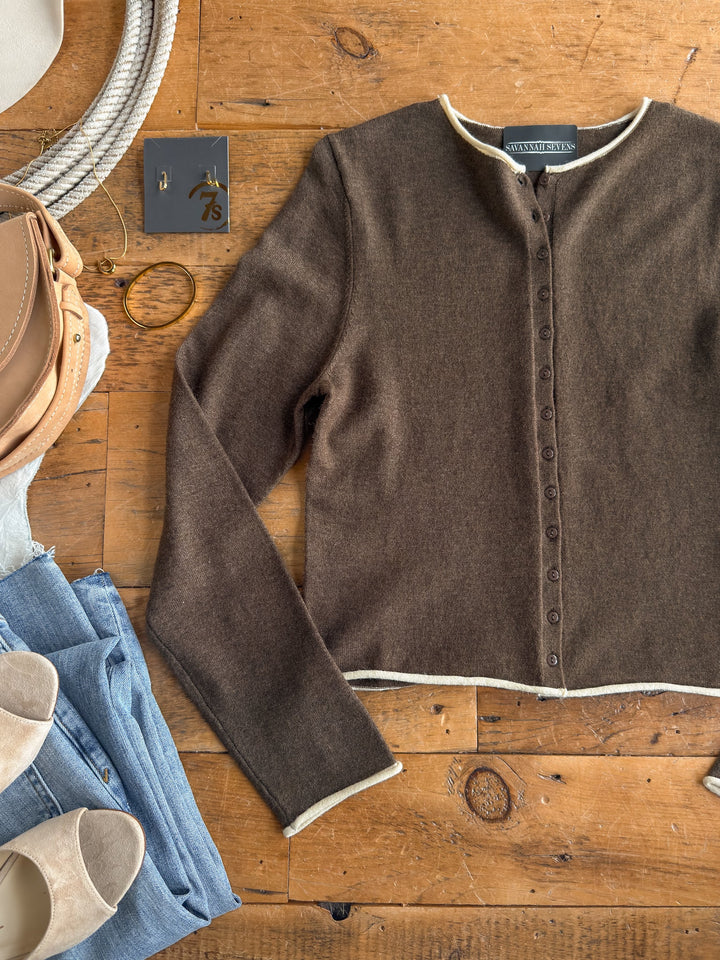 The Camargo Brown Button Up Cardigan from Savannah Sevens western life{&}style is displayed on a wooden surface, complemented by blue jeans, beige shoes, a tan purse, gold hoop earrings, a ring, and a decorative metal plate.