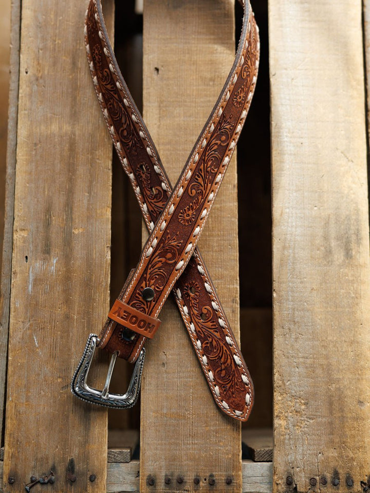 The Camarillo Belt by Savannah Sevens western life{&}style, featuring decorative tooling and a Southwest stamped buckle, rests crossed on a rustic wooden pallet.