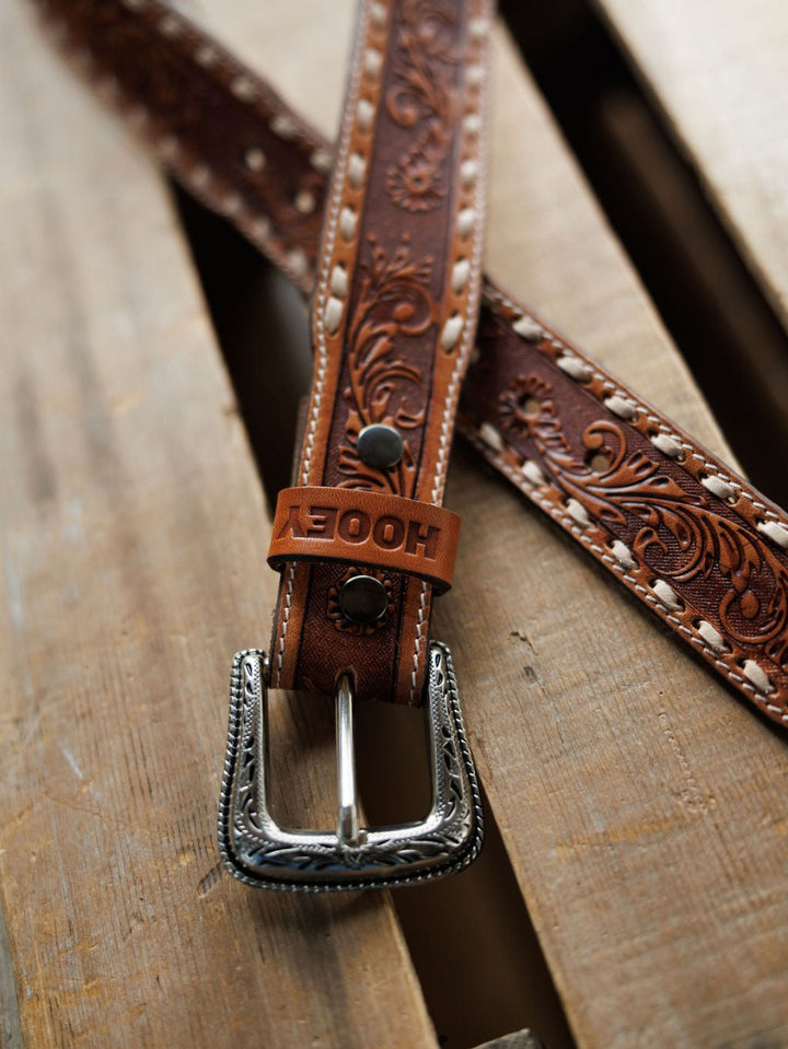 The Savannah Sevens western life{&}style Camarillo Belt, featuring decorative floral embossing, white stitching, a silver buckle, and a tan leather loop stamped with "HOOEY," lies on a wooden surface.