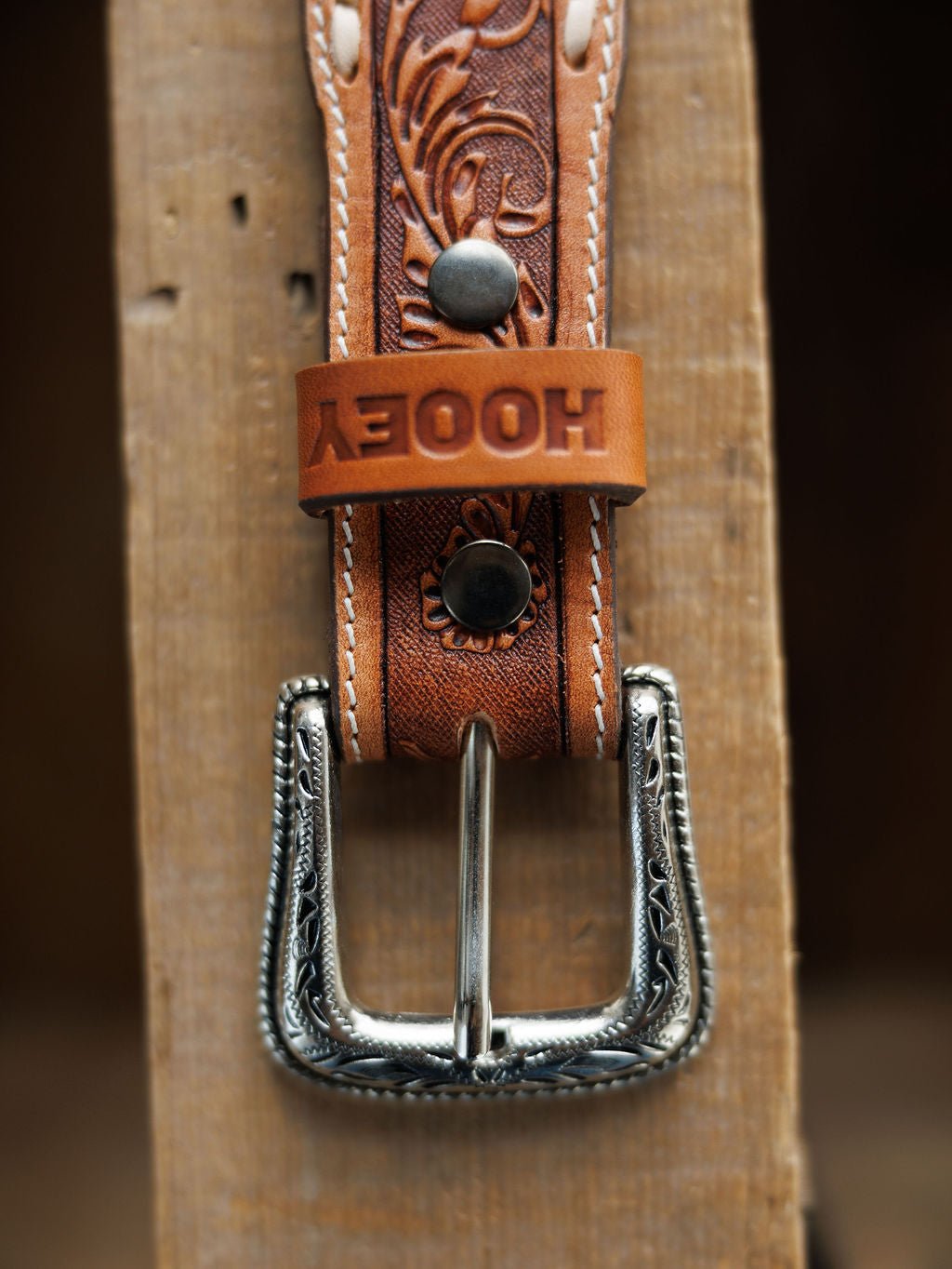 A close-up of the Camarillo Belt by Savannah Sevens western life{&}style, featuring tooled leather, a Southwest-stamped silver buckle, white stitching, and an embossed loop, lying on a wooden surface.