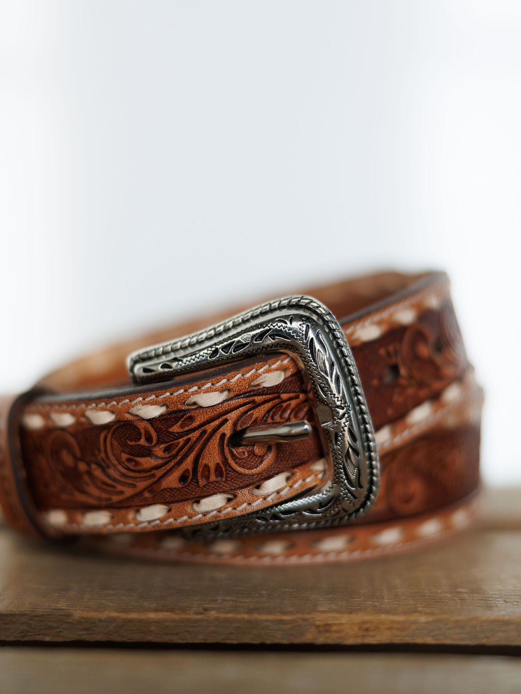 A close-up of the Camarillo Belt by Savannah Sevens western life{&}style, featuring tooled leather with detailed floral patterns, white stitching, and a Southwest stamped buckle, displayed on wood with a blurred background.