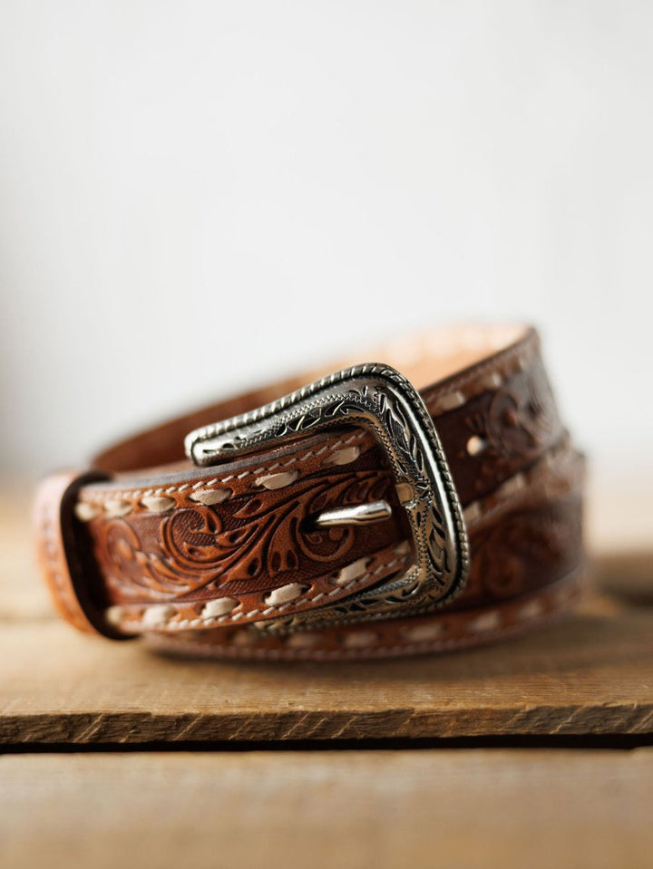 The Camarillo Belt by Savannah Sevens western life{&}style features tooled leather with floral patterns, cream stitching, and a Southwest stamped silver buckle, displayed on a wooden surface.