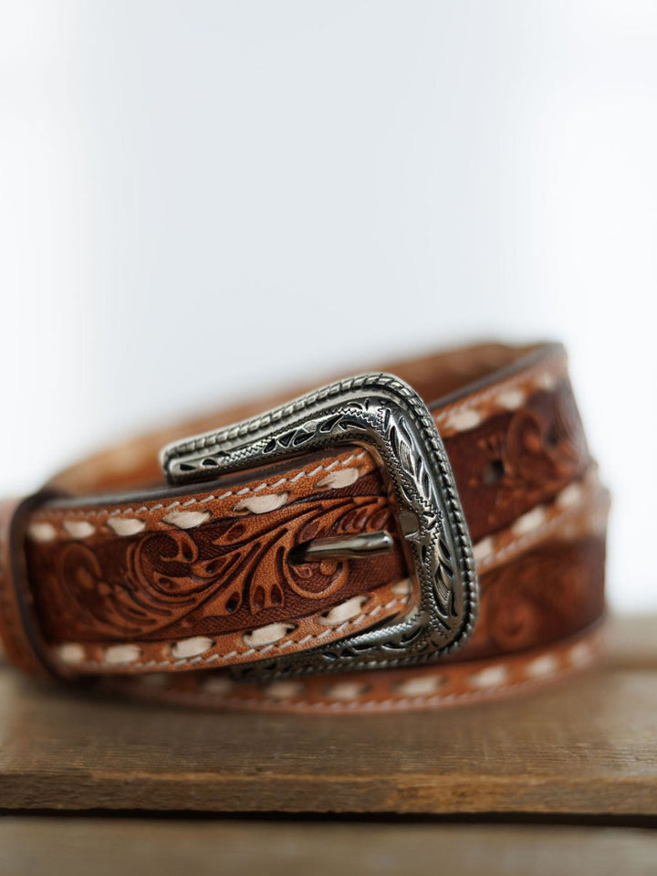 A close-up of the Camarillo Belt by Savannah Sevens western life{&}style, featuring tooled leather with detailed floral patterns, white stitching, and a Southwest stamped buckle, displayed on wood with a blurred background.