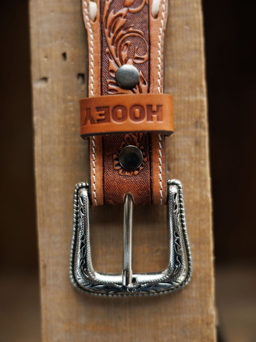 A close-up of the Camarillo Belt by Savannah Sevens western life{&}style, featuring tooled leather, a Southwest-stamped silver buckle, white stitching, and an embossed loop, lying on a wooden surface.