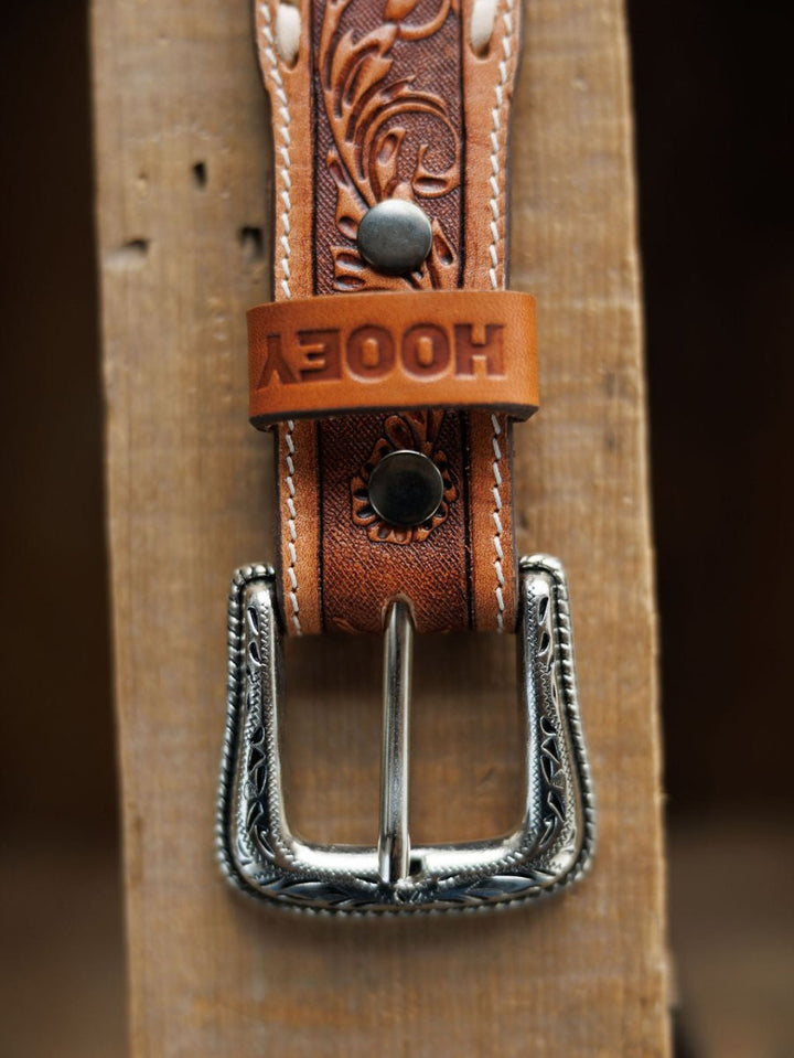A close-up of the Camarillo Belt by Savannah Sevens western life{&}style, featuring tooled leather, a Southwest-stamped silver buckle, white stitching, and an embossed loop, lying on a wooden surface.