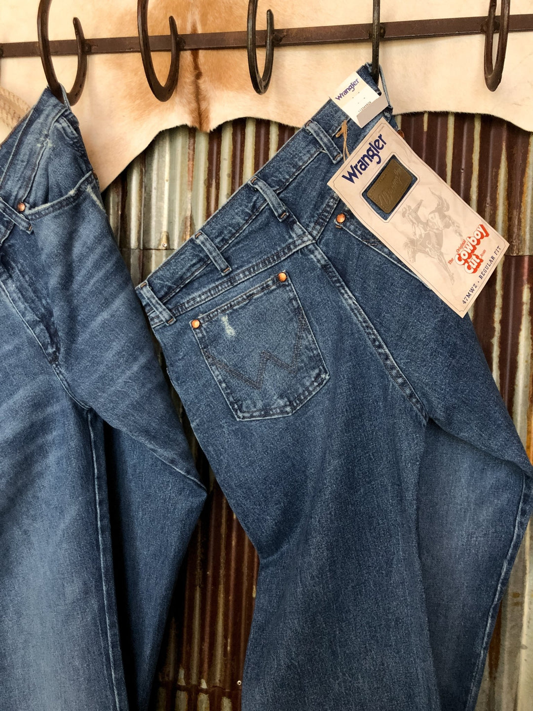 Two pairs of The Camden High Rise 90's Straight Leg Jeans by Savannah Sevens western life{&}style hang on hooks, one with original tags. A corrugated metal wall and tan cowhide are seen in the background.