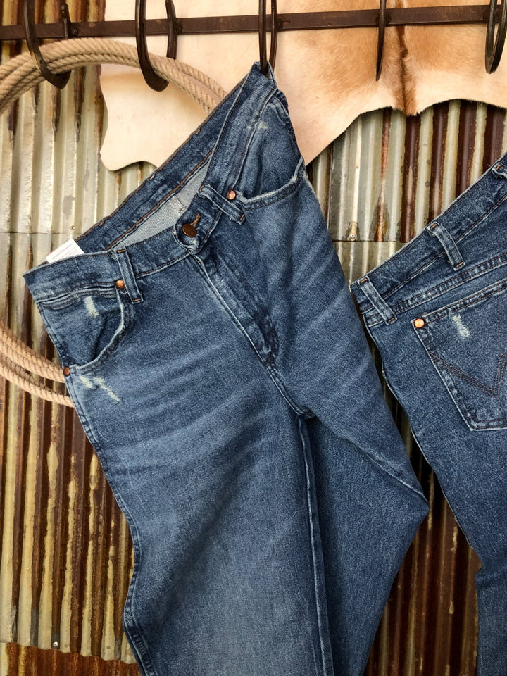 A pair of The Camden High Rise 90's Straight Leg Jeans by Savannah Sevens western life{&}style hangs on a rustic metal rod with rope and cowhide in the background, set against a corrugated metal wall.