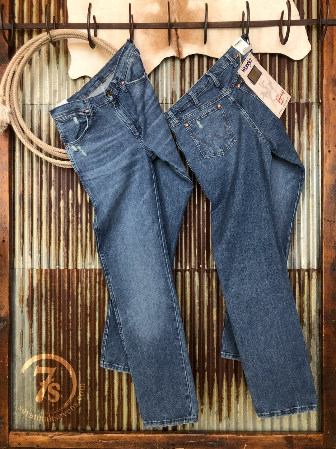 Two pairs of The Camden High Rise 90's Straight Leg Jeans by Savannah Sevens western life{&}style hang on hooks against a rustic metal wall; one pair has tags, both show off classic cowboy fit and timeless style. A rope and cowhide add authentic western flair.