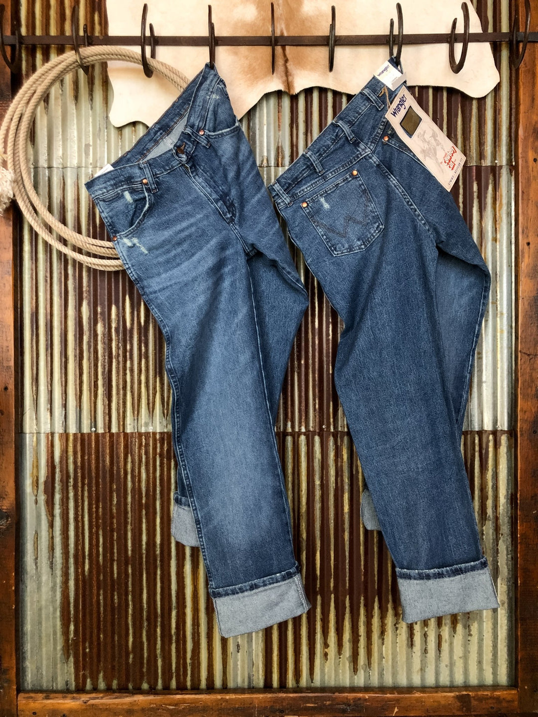 Two pairs of blue jeans, including The Camden High Rise 90's Straight Leg Jean by Savannah Sevens western life{&}style, hang cuffed on hooks against corrugated metal and wood. A coiled rope is in the upper left, and one pair has a tag on its waistband.