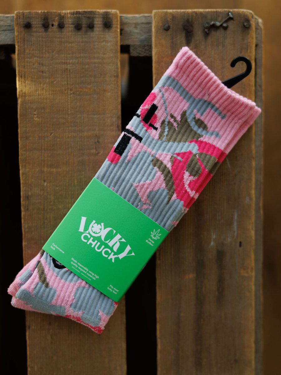 A pair of Camo Crew Socks by Savannah Sevens western life{&}style in pink with black, red, and gray abstract designs and a green Lucky Chuck label is displayed on a wooden surface.