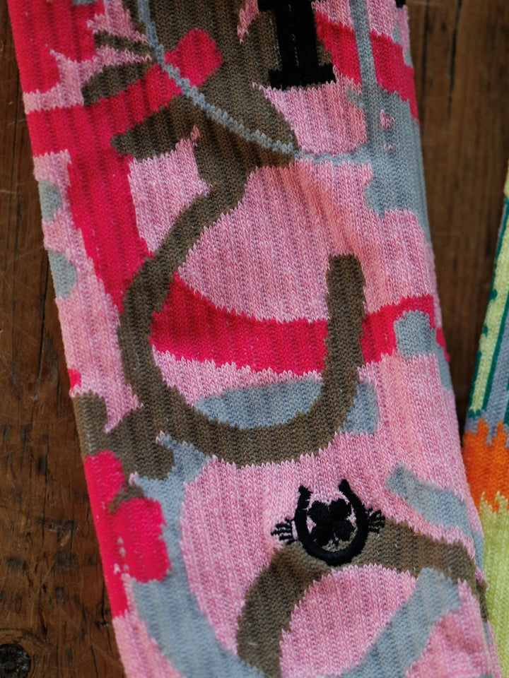 A close-up of the Savannah Sevens western life{&}style Camo Crew Socks shows abstract pink, gray, and brown patterns with a small black smiling face design near the bottom, laid on a wooden surface.