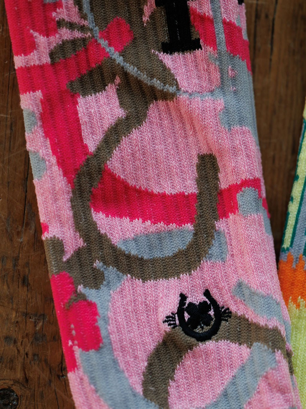 A close-up of the Savannah Sevens western life{&}style Camo Crew Socks shows abstract pink, gray, and brown patterns with a small black smiling face design near the bottom, laid on a wooden surface.
