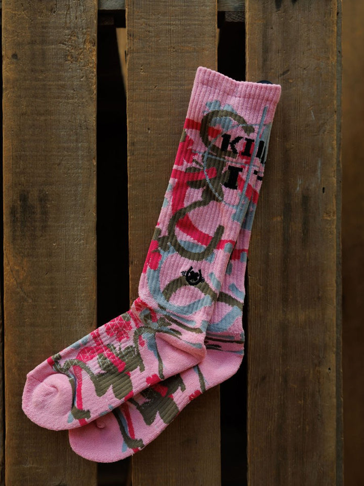 The Camo Crew Socks by Savannah Sevens western life{&}style are pink, breathable crew socks with abstract gray and red graffiti-inspired patterns and black text, shown against a vertical wooden pallet background.