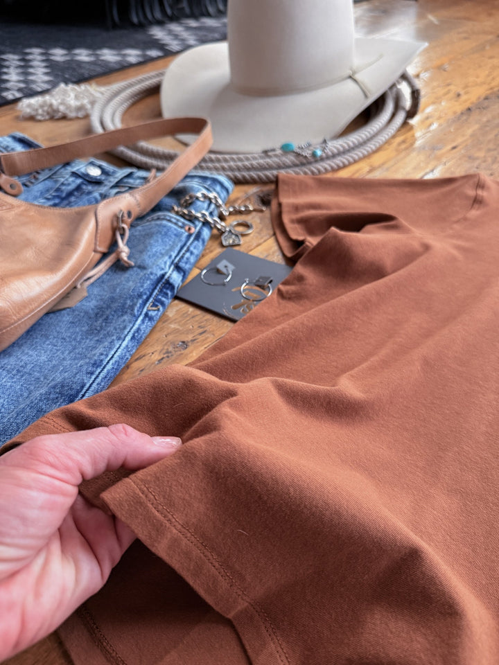 A hand holds the hem of a Campbell Caramel Basic Tee laid on a wooden surface with blue jeans, a tan handbag, a cowboy hat, and silver jewelry.