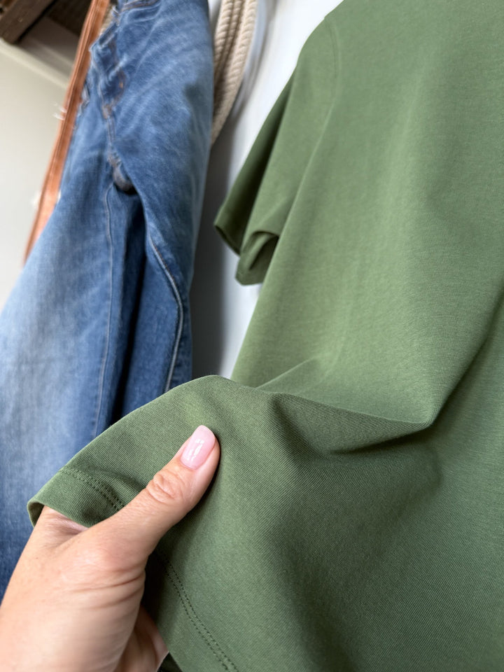 A hand with light pink nails holds the edge of the Campbell Fern Basic Tee by Savannah Sevens western life{&}style in green. In the background, a pair of blue jeans hangs on a hook against a white wall.