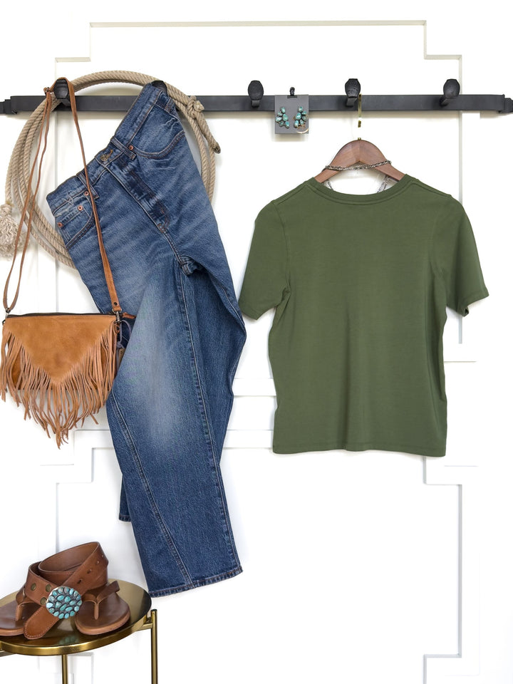The Campbell Fern Basic Tee by Savannah Sevens western life{&}style, featuring a feminine fit and short sleeves in green pima cotton, is styled with blue jeans, a brown fringe purse, brown sandals, and turquoise earrings—all neatly arranged against a white wall with a black hook rack.