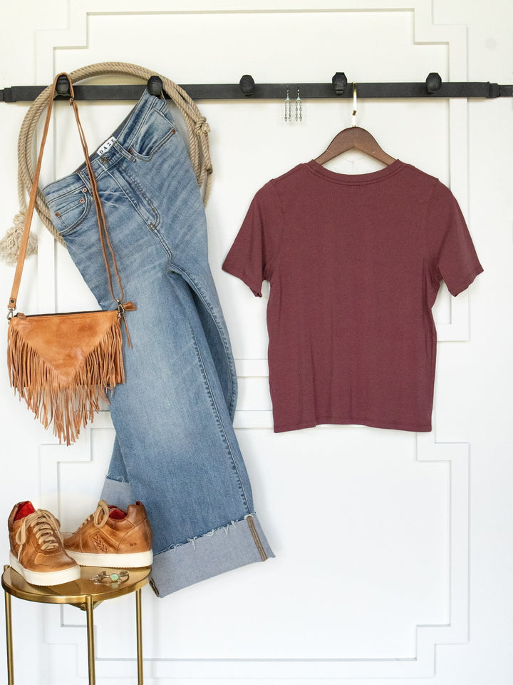 A casual outfit featuring light blue jeans, the Campbell Rosewood Basic Tee by Savannah Sevens western life{&}style in maroon pima cotton, a tan fringe purse, tan sneakers with red accents, and a rope belt, all displayed on a white wall with a black hook rack.