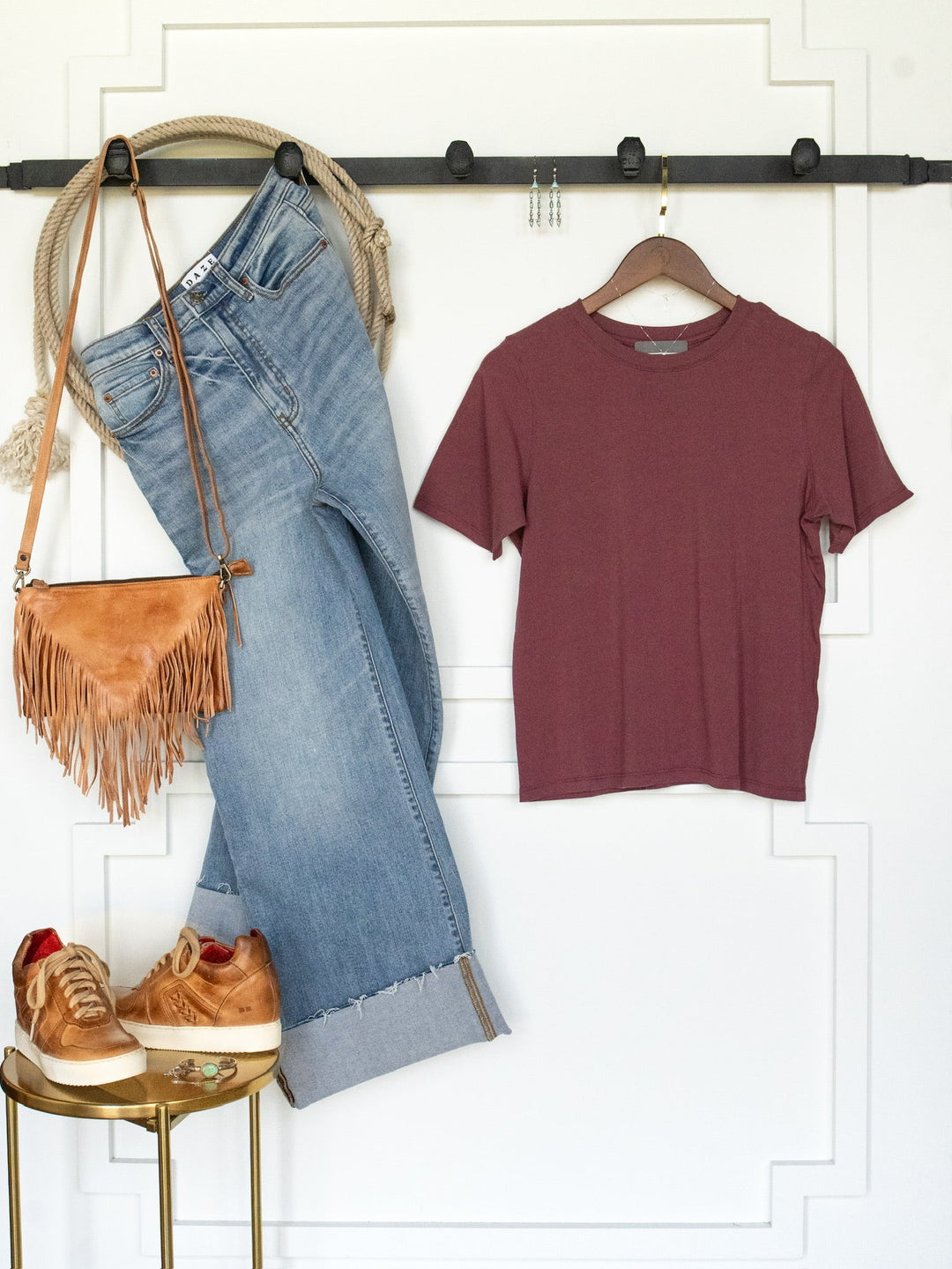 A casual outfit hung on the wall features light blue jeans, the Campbell Rosewood Basic Tee from Savannah Sevens western life{&}style—a maroon short sleeve pima cotton tee with a feminine fit—paired with a tan fringe purse, gold sneakers, a rope belt, jewelry, and rings displayed on a small round table.