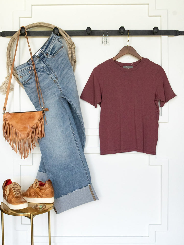 A casual outfit hung on the wall features light blue jeans, the Campbell Rosewood Basic Tee from Savannah Sevens western life{&}style—a maroon short sleeve pima cotton tee with a feminine fit—paired with a tan fringe purse, gold sneakers, a rope belt, jewelry, and rings displayed on a small round table.