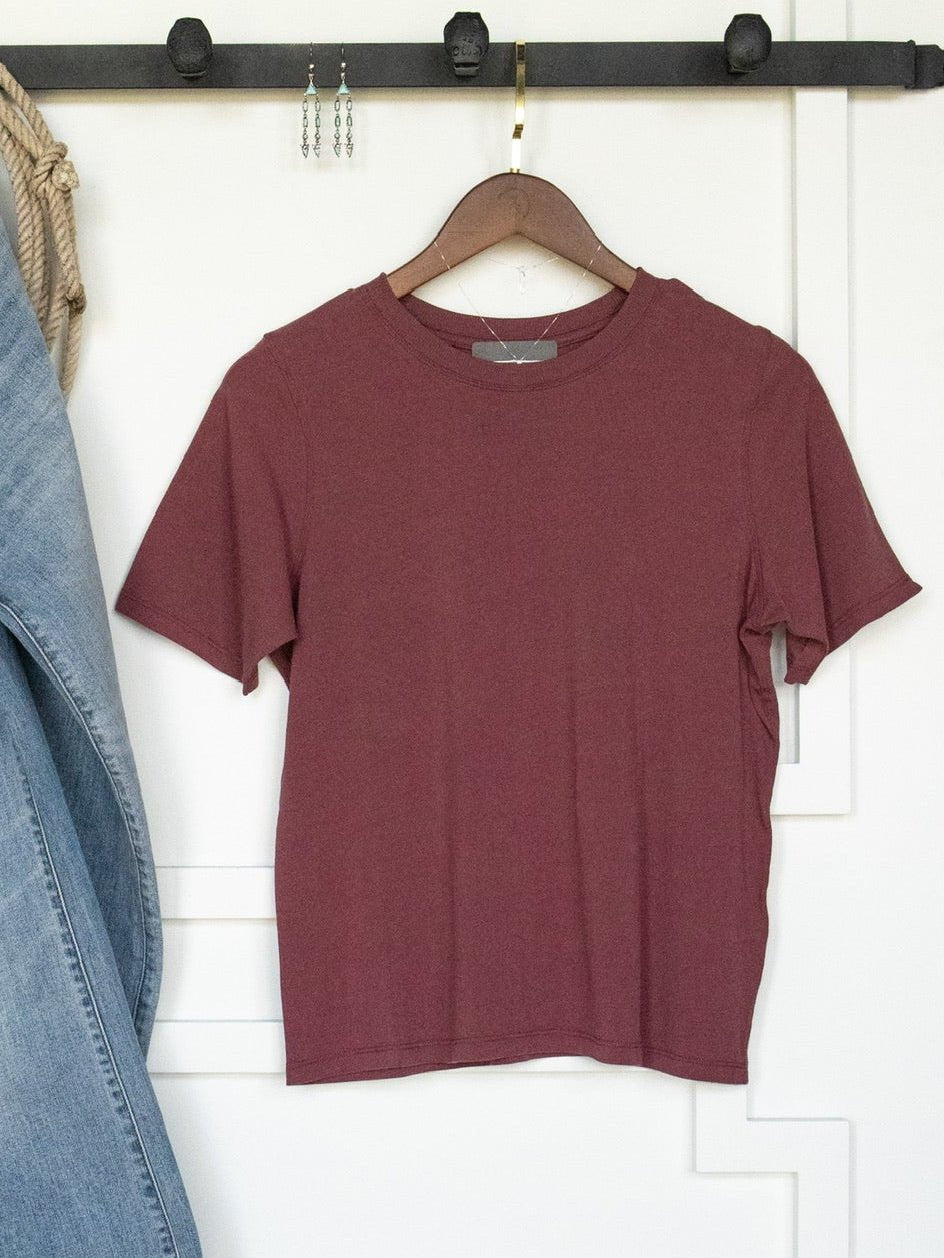 The Campbell Rosewood Basic Tee by Savannah Sevens western life{&}style, a plain maroon short sleeve pima cotton shirt with a feminine fit, is displayed on a wooden hanger suspended from a black wall hook rail. A denim jacket and rope bag are partially visible to the left.