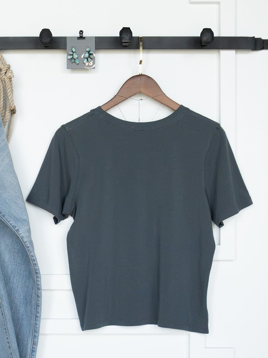 The Campbell Slate Basic Tee by Savannah Sevens western life{&}style, a plain dark gray pima cotton top with a feminine fit, hangs on a wooden hanger against a white wall, accompanied by a denim jacket partly visible on the left and a pair of earrings displayed above.