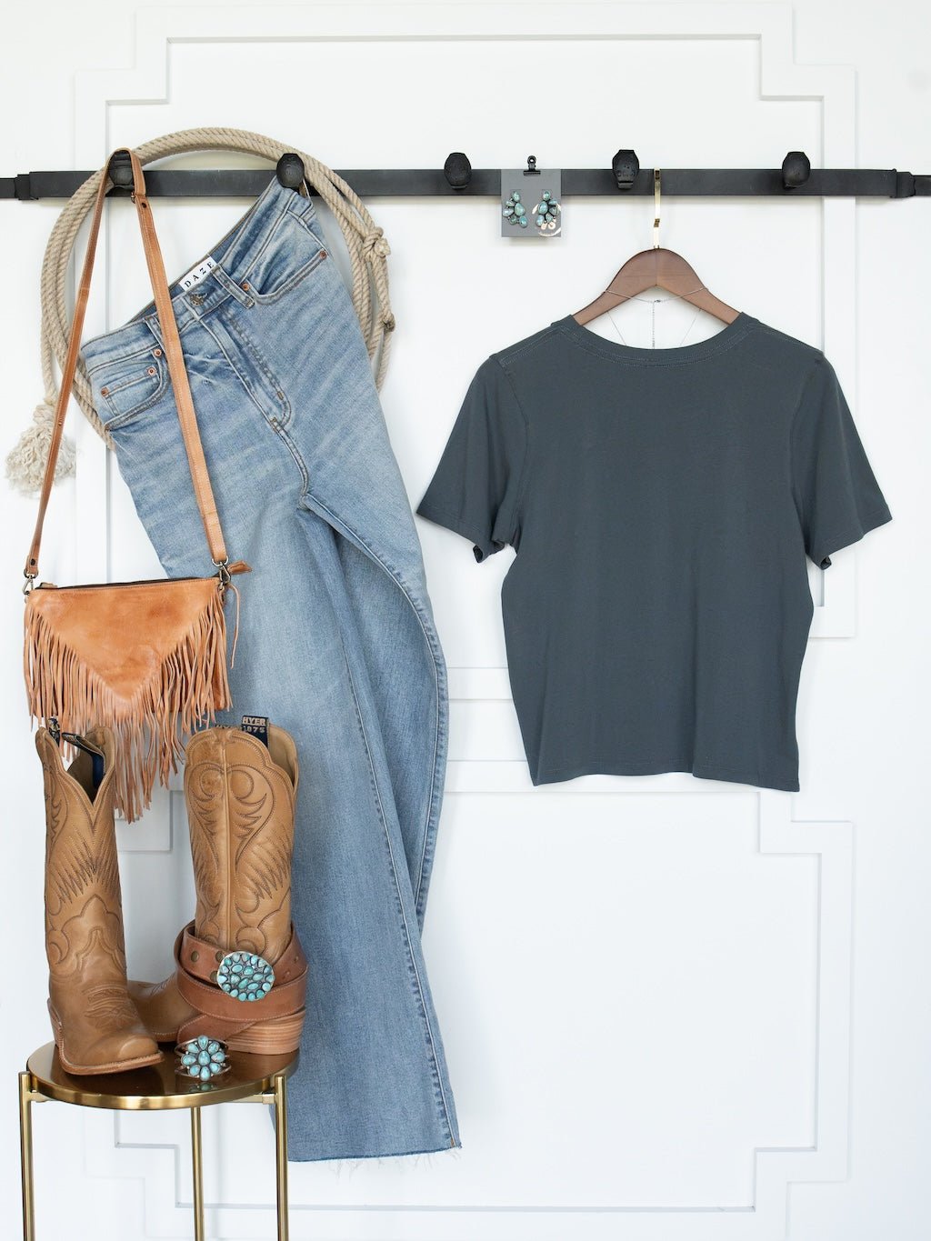 The Campbell Slate Basic Tee by Savannah Sevens western life{&}style, a pair of jeans, tan cowboy boots with turquoise accents, a tan fringed purse, and earrings are neatly arranged on a wall rack and stool.