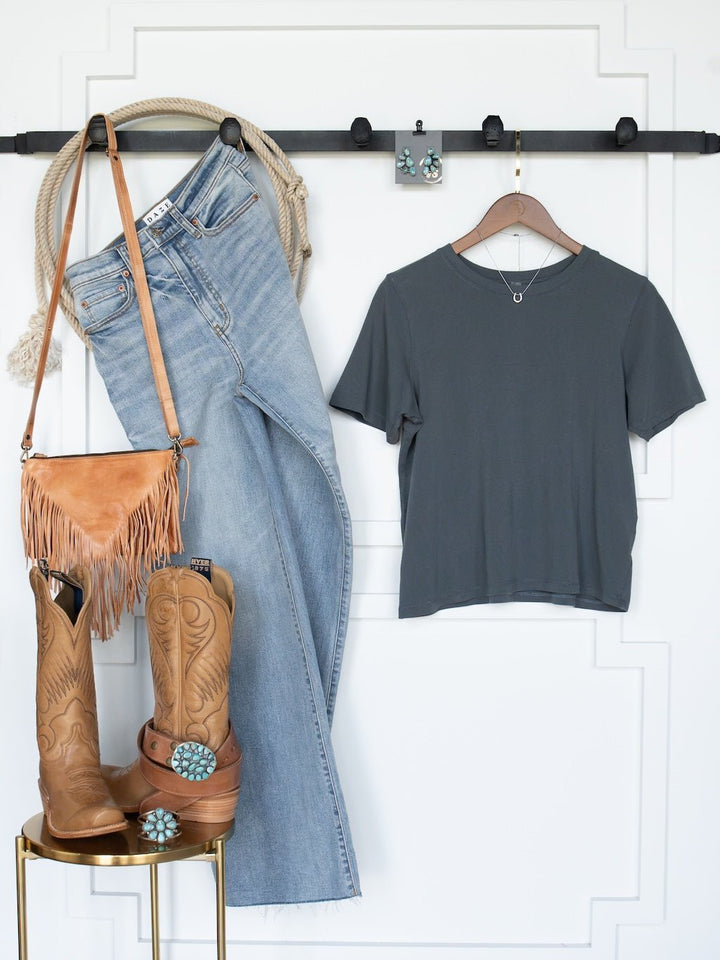 A casual outfit featuring the Campbell Slate Basic Tee by Savannah Sevens western life{&}style—a gray short sleeve pima cotton tee—paired with light blue jeans, cowboy boots, a tan fringe purse, a rope, and turquoise jewelry, all displayed neatly on a white wall and set atop a gold stool.