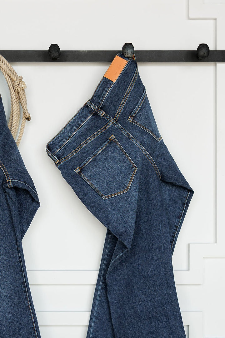 A pair of The Camryn Premier Rise Slim Straight Leg Jeans by Savannah Sevens western life{&}style hangs folded over a black wall hook, with a white paneled wall and part of a rope visible in the background.