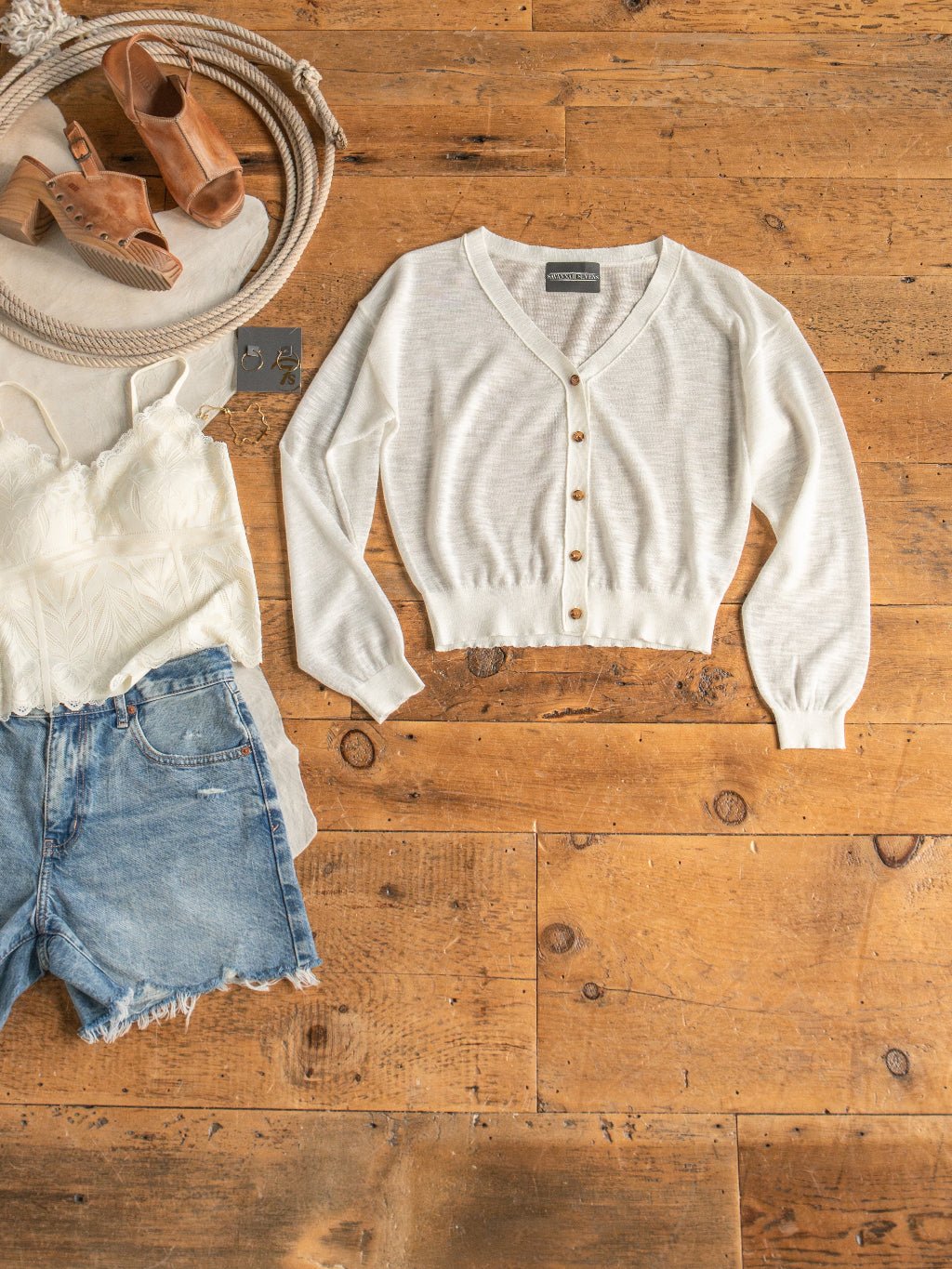 A flat lay on a wooden floor featuring the Caney White Button Up Cardigan by Savannah Sevens western life{&}style, a white lace camisole, denim shorts, tan heeled sandals, a beige bag, and a coiled rope accessory.