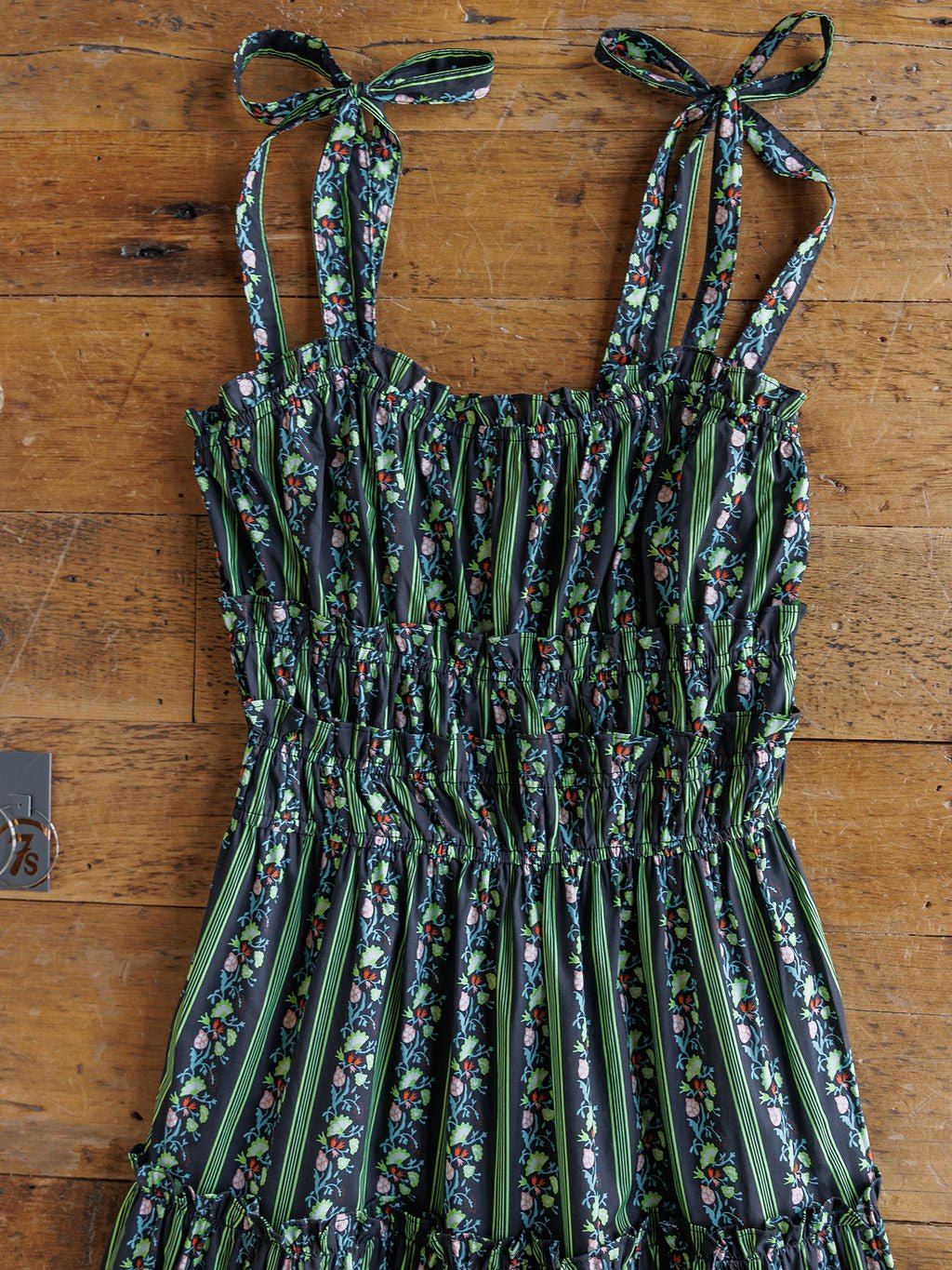 The Canova Green Floral Dress by Savannah Sevens western life{&}style features a smocked bodice, adjustable tie straps, and a green floral print on a dark background, displayed flat on a wooden surface.