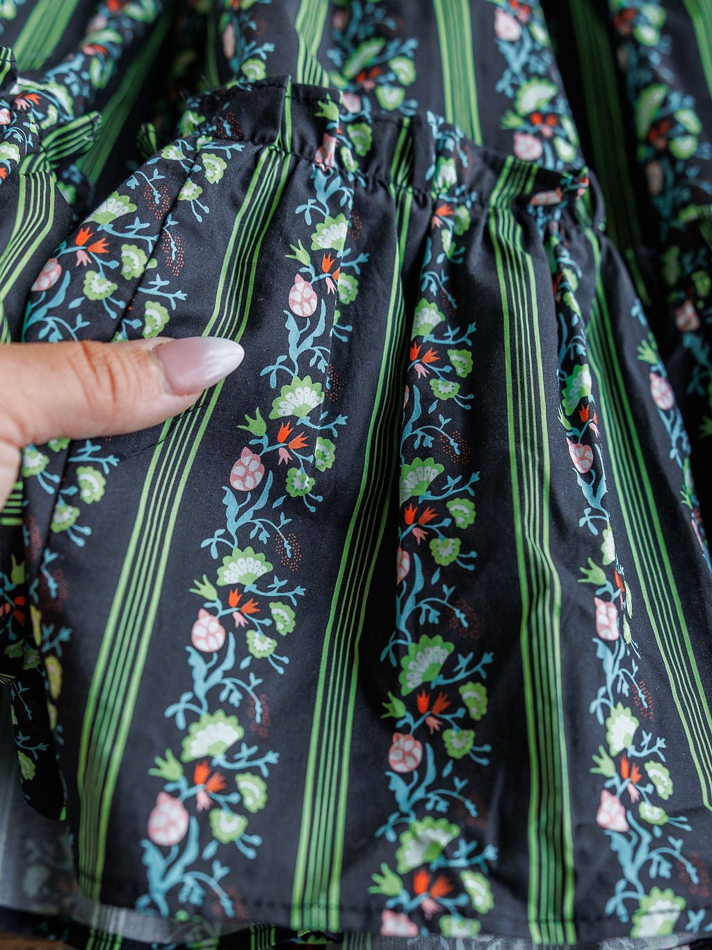 A close-up of a hand holding the pocket of the Canova Green Floral Dress by Savannah Sevens western life{&}style, featuring black cotton fabric with vertical green stripes and a floral pattern in pink, red, and green.