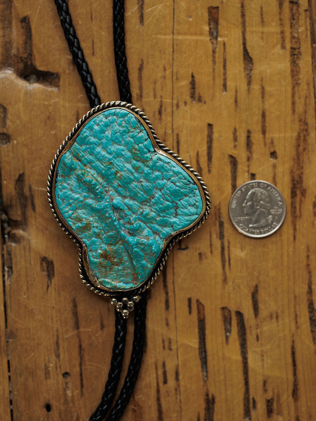 The Carlsbad Bolo by Savannah Sevens western life{&}style, featuring a large, irregular turquoise stone with silver edging, is shown next to a U.S. quarter on wood for size reference.