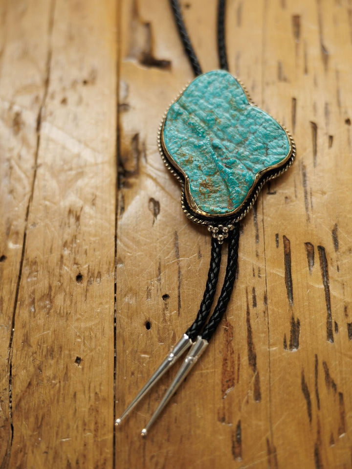 The Carlsbad Bolo by Savannah Sevens western life{&}style features a turquoise slide, braided black leather cords, and silver tips. This handmade piece pairs classic style with modern elegance on a rustic wooden backdrop.