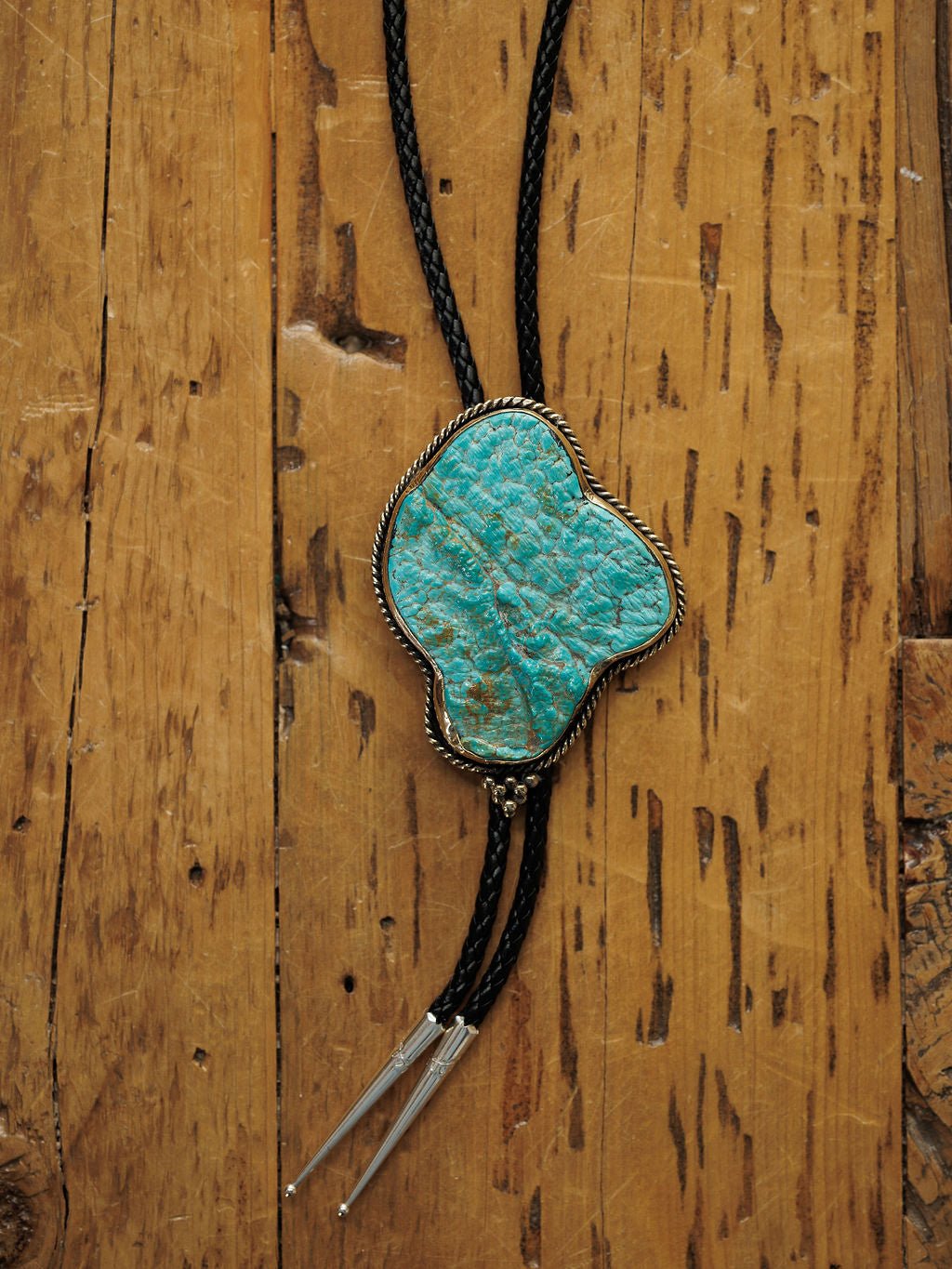 The Carlsbad Bolo by Savannah Sevens western life{&}style, featuring turquoise in a silver setting and a black braided cord, is displayed on a rustic wooden surface.