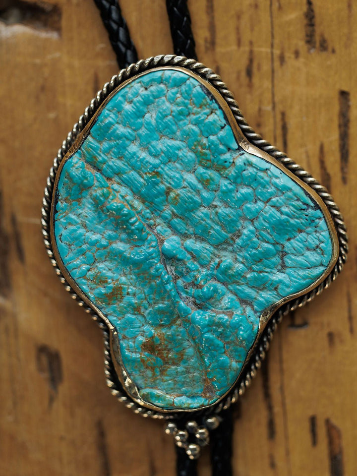 A close-up of the Carlsbad Bolo by Savannah Sevens western life{&}style, featuring a turquoise stone in decorative silver on a black braided cord, displayed against a wooden background.