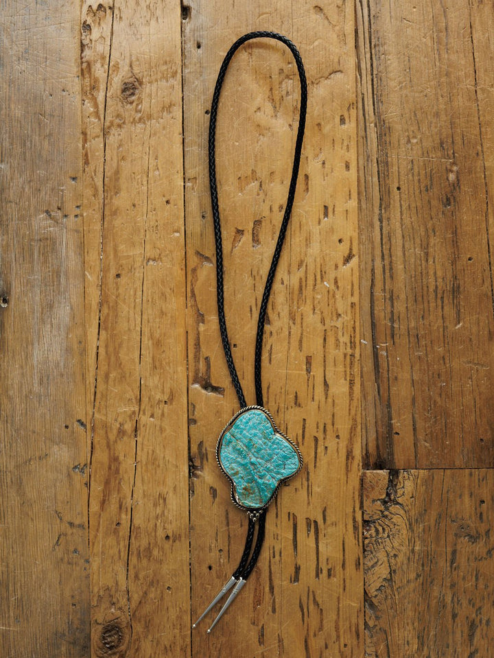 The Carlsbad Bolo by Savannah Sevens western life{&}style, featuring a turquoise accent, black braided cord, and silver tips, rests on rustic wood—highlighting its striking handmade design.