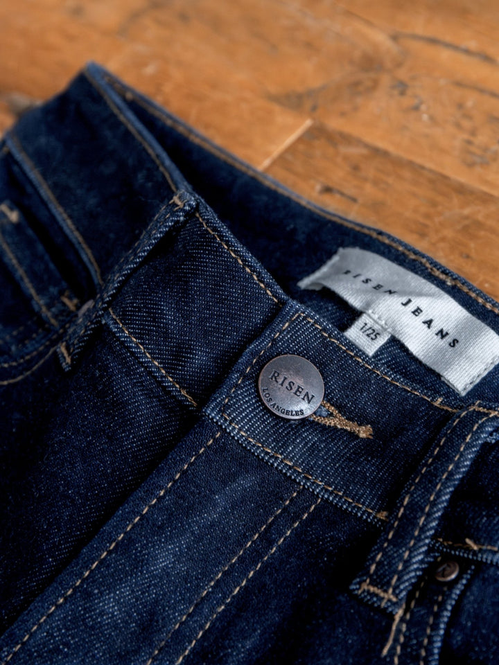 A close-up of The Carson High Rise Wide Leg Jean by Savannah Sevens western life{&}style, featuring dark wash denim, visible stitching, a metal button, and a fabric label with size 12/31, displayed on a wooden surface.